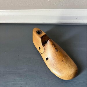 May include: A vintage wooden shoe last, light brown in color, with a rounded toe and heel. The shoe form has a metal insert at the top and a series of small holes and markings. The shoe last is designed to shape and mold shoes.