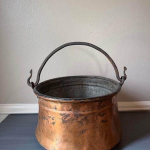 May include: A vintage copper cauldron with a dark metal handle. The cauldron has a wide, rounded body and a slightly flared rim. The copper has a rich, aged patina, with areas of darker oxidation. The handle is attached with curved hooks.