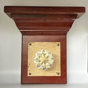 May include: A decorative wooden shelf with a dark red finish. It features a square, gold-colored metal plate with an ornate design and a clear, star-shaped glass knob. The shelf is mounted with screws.