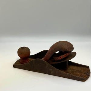May include: A vintage metal hand plane featuring a dark, weathered iron body with visible oxidation. It includes a rounded wooden front knob and a curved, red-painted metal handle. The internal mechanism shows a circular red adjustment wheel.