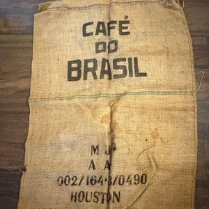 May include: A rectangular tan burlap coffee sack featuring the black printed text CAFÉ DO BRASIL, followed by the letters M J A A, the numerical sequence 002/164-8/0490, and the word HOUSTON. The woven fabric has a coarse texture and visible seams.