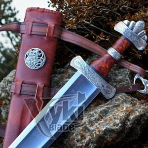 Könnte beinhalten: Ein Wikinger-Schwert mit polierter Stahlklinge und kunstvollem silberfarbenem Knauf. Der Griff ist rotbraun. Das Schwert wird mit einer braunen Lederscheide und Riemen präsentiert. Das Wort "Blade" ist auf der Klinge zu sehen.