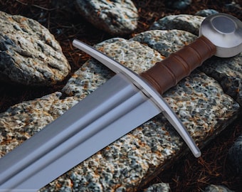 May include: A medieval sword with a silver blade and a brown leather-wrapped handle. The sword features a rounded pommel and a crossguard. The sword is resting on a bed of rocks and earth, suggesting an outdoor setting.