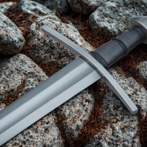Op de afbeelding: Een middeleeuws zwaard met een zilveren lemmet en pareerstang. De handgreep is gewikkeld in donkergrijs materiaal en de pommel is een afgeronde, zilveren dop. Het zwaard rust op een bed van rotsen en droog gebladerte.