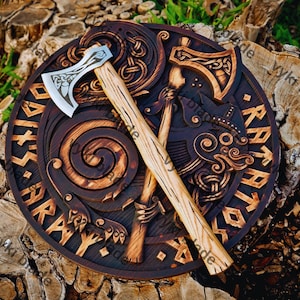 May include: A round, dark brown wooden wall hanging with intricate carvings of Viking symbols and an axe. The axe has a silver blade and a light brown wooden handle. The wall hanging has the text "Ragnar" carved into it.