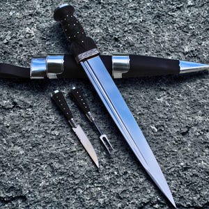 May include: A Scottish dirk with a polished steel blade and a black handle with silver accents. The dirk is accompanied by a matching sheath and two smaller knives. The set is displayed on a textured gray surface.