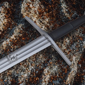 Puede incluir: Una espada de estilo medieval con una hoja y una guarda plateadas, un mango negro y un pomo redondeado. La espada descansa sobre una superficie rocosa y texturizada. La hoja tiene el texto "Vki Blade".