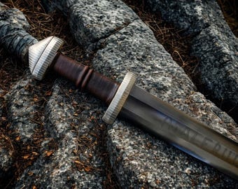 May include: A detailed image of a Viking sword resting on a stone surface. The sword features a dark, weathered blade with the inscription "WAVERIND". The handle is wrapped in a textured, gray material, and the hilt is silver.