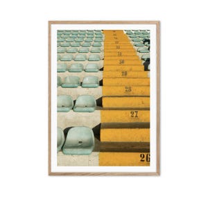 Könnte beinhalten: Gerahmter Druck mit einer Stadionbestuhlung. Reihen hellblauer Sitze stehen im Kontrast zu leuchtend gelben Stufen, die jeweils mit schwarzen Zahlen markiert sind. Die Komposition erzeugt einen auffälligen Kontrast von Farben und Texturen.