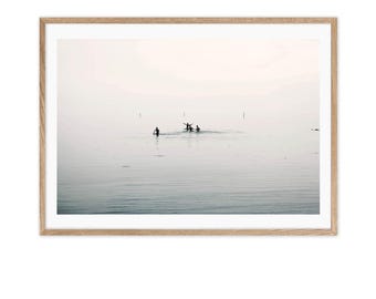 May include: A black and white photograph of four people in the water, silhouetted against a hazy, light pink sky. The water is calm and still, and the sky is a soft, muted pink.