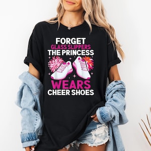 K&ouml;nnte beinhalten: Schwarzes T-Shirt mit einem pinkfarbenen Aufdruck, auf dem steht "Vergiss Glaspantoffeln, die Prinzessin tr&auml;gt Cheerleader-Schuhe". Der Aufdruck zeigt auch ein Paar pinkfarbene Cheerleader-Schuhe und einen pinkfarbenen Pompon.