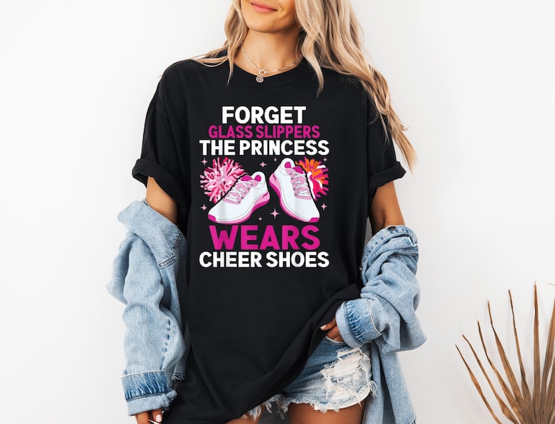K&ouml;nnte beinhalten: Schwarzes T-Shirt mit einem pinkfarbenen Aufdruck, auf dem steht "Vergiss Glaspantoffeln, die Prinzessin tr&auml;gt Cheerleader-Schuhe". Der Aufdruck zeigt auch ein Paar pinkfarbene Cheerleader-Schuhe und einen pinkfarbenen Pompon.