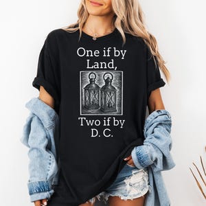 May include: Black t-shirt with the text "One if by Land, Two if by D.C." and a graphic of two lanterns. The shirt is a dark color and has a casual style.