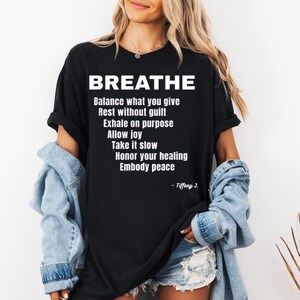 May include: Black t-shirt with the word "BREATHE" in large white letters. Below, a list of affirmations is printed in white. The shirt is worn with a light blue denim jacket.