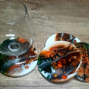 May include: A clear glass wine glass rests on a colorful coaster. The coasters feature abstract designs with orange, brown, green, and white hues. Additional coasters with similar patterns are visible.