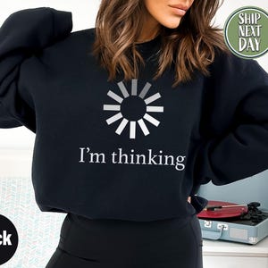 Pode incluir: Camisola preta com um gráfico branco de um símbolo de carregamento e o texto "I'm thinking". A camisola é de cor preta lisa. O desenho está centrado na frente da peça.