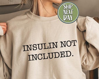 May include: A sand-colored sweatshirt with the text "INSULIN NOT INCLUDED" in black. The sweatshirt has long sleeves and a crew neck. A green circle with the words "SHIP NEXT DAY" is visible.