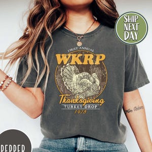 May include: Dark gray t-shirt with a distressed graphic featuring the text "WKRK Thanksgiving Turkey Drop 1978" and a turkey illustration. The shirt is a vintage style and is paired with blue jeans and white sneakers.