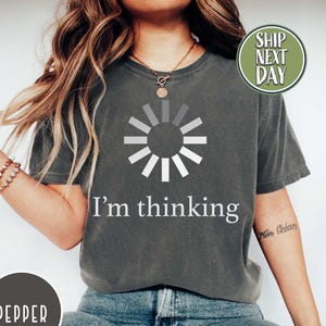 May include: Pepper-colored t-shirt with a white loading symbol and the text "I'm thinking." The shirt is paired with blue jeans and white sneakers. A circular label reads "Pepper."