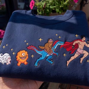 Pode incluir: Camisola azul marinho com personagens de desenhos animados bordados. Apresenta um super-her&oacute;i correndo, um urso montando um dinossauro, um monstro e uma mulher gritando. Acentos de estrelas douradas tamb&eacute;m s&atilde;o bordados na camisola.