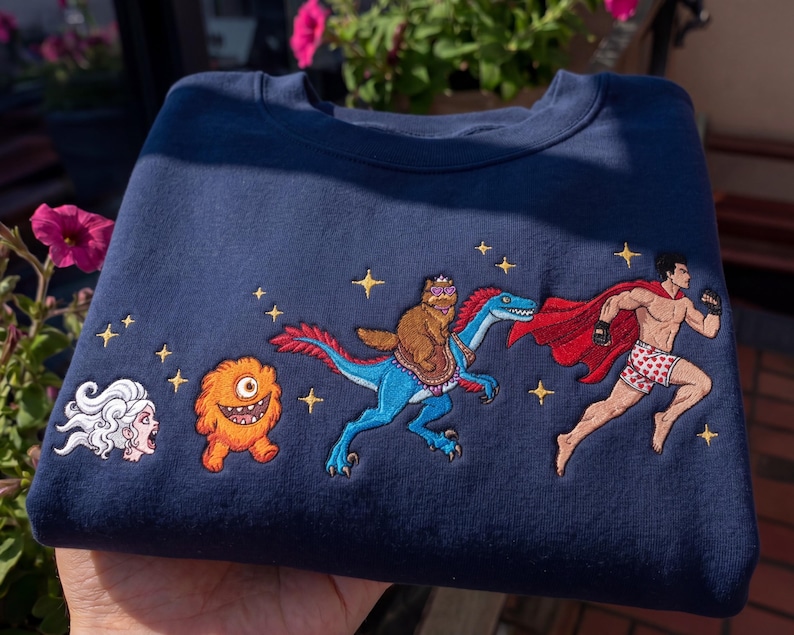 Pode incluir: Camisola azul marinho com personagens de desenhos animados bordados. Apresenta um super-her&oacute;i correndo, um urso montando um dinossauro, um monstro e uma mulher gritando. Acentos de estrelas douradas tamb&eacute;m s&atilde;o bordados na camisola.