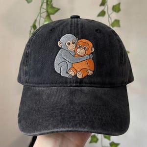 May include: Black baseball cap with an embroidered design of two hugging monkeys. One monkey is gray, and the other is orange. The cap has a curved brim and an adjustable strap.