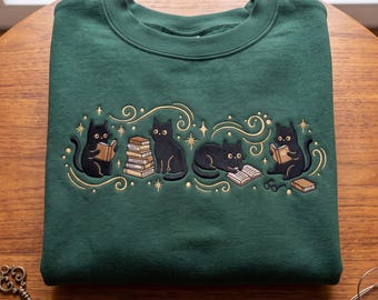 Könnte beinhalten: Ein waldgrüner Rundhals-Pullover mit einer Stickerei von vier schwarzen Katzen, die mit Bücherstapeln und offenen Büchern interagieren, akzentuiert durch goldfarbene geschwungene Linien und kleine Sternformen auf der Brust.
