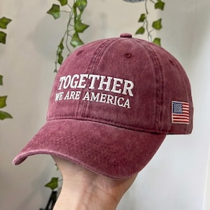 May include: A maroon baseball cap with the text "TOGETHER WE ARE AMERICA" in white. An American flag patch is on the side. The cap is made of a soft, worn material.