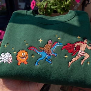 Pode incluir: Um moletom verde escuro com personagens de desenhos animados bordados. O design inclui uma figura de cabelo branco, um monstro laranja, um urso montado em um dinossauro azul e um super-her&oacute;i. Estrelas douradas est&atilde;o espalhadas.