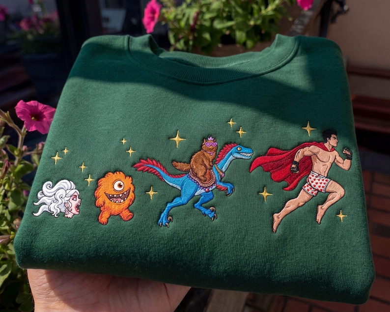 Pode incluir: Um moletom verde escuro com personagens de desenhos animados bordados. O design inclui uma figura de cabelo branco, um monstro laranja, um urso montado em um dinossauro azul e um super-her&oacute;i. Estrelas douradas est&atilde;o espalhadas.