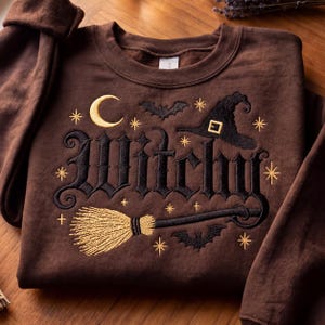 May include: A dark brown crewneck sweatshirt featuring black and gold embroidery. The design includes the word Witchy in a gothic font, a gold crescent moon, a black witch hat, a gold broom, two black bats, and several small gold stars.