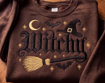 May include: A dark brown crewneck sweatshirt featuring black and gold embroidery. The design includes the word Witchy in a gothic font, a gold crescent moon, a black witch hat, a gold broom, two black bats, and several small gold stars.