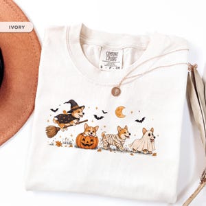 May include: An ivory t-shirt featuring a graphic of four Corgi dogs in Halloween costumes. One Corgi wears a witch hat and cape on a broom, one sits in a pumpkin, one is wrapped like a mummy, and one wears a ghost sheet. Bats, stars, and a moon are included.