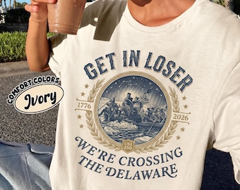 May include: Ivory-colored t-shirt with the text "GET IN LOSER" and "WE'RE CROSSING THE DELAWARE". The design features a circular illustration of a historical scene, with the years 1776 and 2026. The shirt also has the words "COMFORT COLORS" and "USA 250".