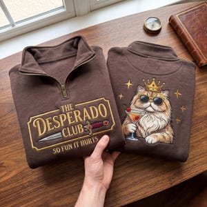 May include: Two brown sweatshirts with a quarter-zip collar. One features "The Desperado Club" text with a dagger graphic. The other has a cat wearing sunglasses and a crown, holding a cocktail. Both have embroidered designs.