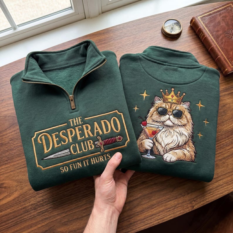 May include: Two dark green sweatshirts with a quarter-zip collar. One features "The Desperado Club" text with a sword graphic. The other has a cat wearing sunglasses and a crown, holding a cocktail. Both are embroidered.