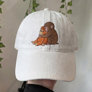 May include: White baseball cap with a curved brim. The cap features an embroidered design of two orangutans hugging. One orangutan is brown, and the other is orange. The cap is made of a soft, durable fabric.