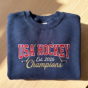 May include: Navy blue sweatshirt with embroidered text "USA HOCKEY Est. 2026 Champions" in red and gold. Features two crossed hockey sticks. The sweatshirt is folded neatly on a light brown surface.
