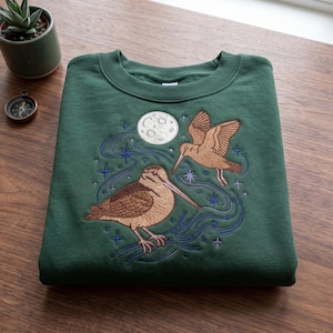 May include: A dark green sweatshirt featuring an embroidered design of two birds, a full moon, and stars. The birds are brown with long beaks, and the design includes blue and purple accents. The sweatshirt is folded on a wooden surface.
