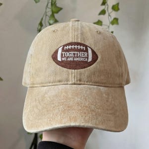 May include: Tan baseball cap with a brown football graphic. The football graphic has the words "TOGETHER WE ARE AMERICA" in white. The cap is made of a soft material and has a curved brim.