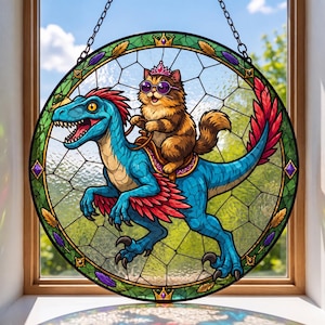 May include: A circular stained glass suncatcher featuring a fluffy brown cat wearing a pink crown and purple sunglasses, riding a blue feathered raptor. The design is enclosed in a green and gold border with a chain for hanging in a window.