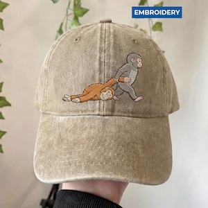 May include: A tan baseball cap with an embroidered design of two monkeys. One monkey is pulling the other, who is lying on the ground. The word "EMBROIDERY" is printed on the top right of the cap.