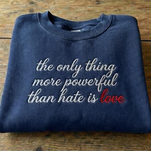 May include: Navy blue sweatshirt with the embroidered quote, "the only thing more powerful than hate is love." The word "love" is in red. The sweatshirt is folded on a wooden surface.