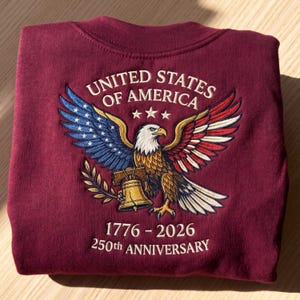 May include: Burgundy sweatshirt featuring an embroidered design of an eagle with outstretched wings, the American flag, and a bell. The text reads "UNITED STATES OF AMERICA" and "1776-2026 250th ANNIVERSARY".