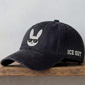 May include: A dark blue baseball cap with a white embroidered bunny wearing sunglasses and a star. The text "ICE OUT" is embroidered on the side of the cap. The cap is sitting on a wooden surface.
