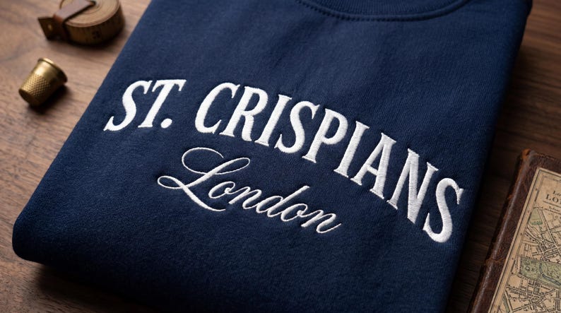 Może przedstawiać: Granatowa bluza z białym haftowanym napisem ST. CRISPIANS pogrubioną czcionką szeryfową oraz słowem London poniżej, zapisanym białą kursywą. Odzież jest złożona i wyeksponowana na drewnianej powierzchni.