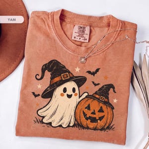 May include: A terracotta-colored crew neck t-shirt featuring a graphic print of a smiling ghost and a jack-o'-lantern, both wearing black witch hats. The design includes small bats and stars. The shirt has a Comfort Colors brand label at the neck.