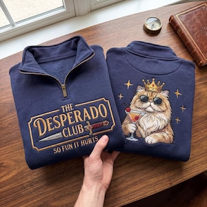 May include: Two navy blue sweatshirts with a mock neck and a zipper. One sweatshirt has the text "THE DESPERADO CLUB SO FUN IT HURTS" embroidered in gold. The other has a cat wearing sunglasses and a crown, holding a cocktail.