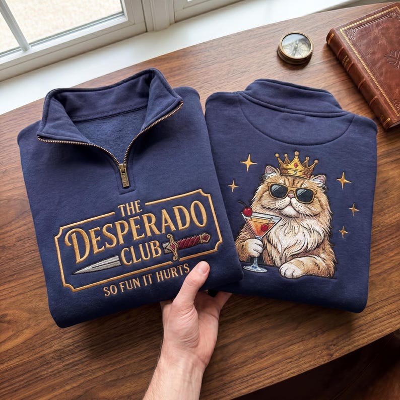 May include: Two navy blue sweatshirts with a mock neck and a zipper. One sweatshirt has the text "THE DESPERADO CLUB SO FUN IT HURTS" embroidered in gold. The other has a cat wearing sunglasses and a crown, holding a cocktail.