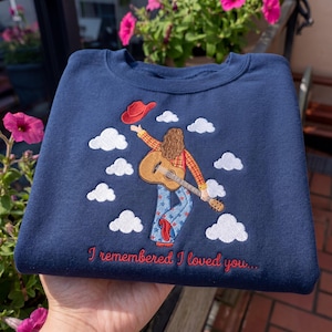 May include: A navy blue sweatshirt featuring an embroidered design of a person with long hair wearing a plaid shirt and star-patterned jeans, holding a guitar and a red cowboy hat, surrounded by white clouds and the text I remembered I loved you.
